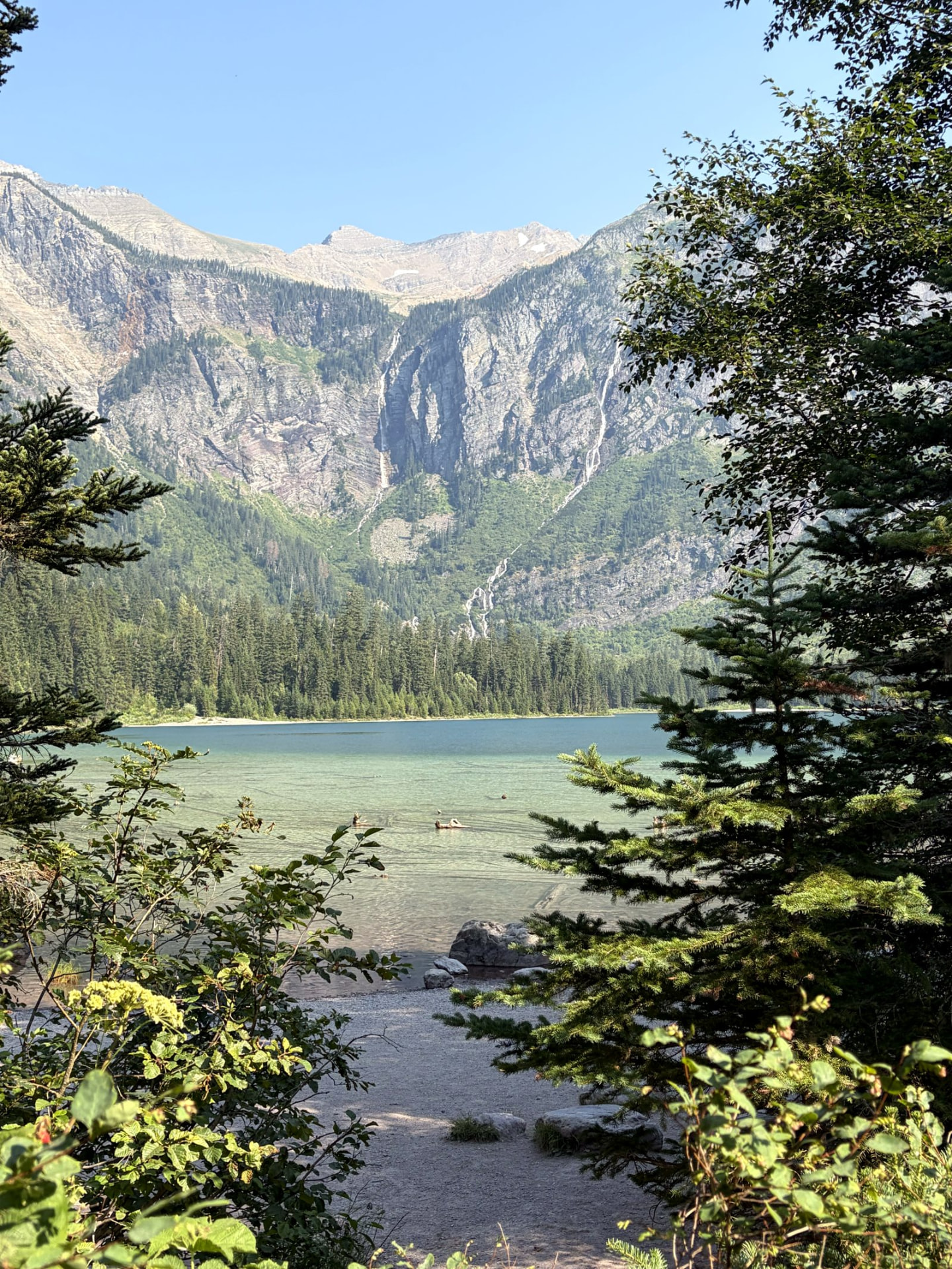 A turquoise mountain lake framed by evergreen branches, with forested slopes, waterfalls and rocky peaks under a clear blue sky.